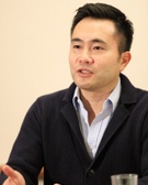 A man speaking during a meeting, dressed in a dark blazer over a blue shirt.