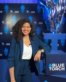 A woman in a blue suit is smiling while standing at a podium with the 'Blue Torch' logo.