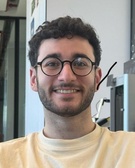 A young man with curly hair and glasses, smiling in an indoor environment.