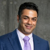 A professional headshot of a smiling man in a blue suit with a purple tie.