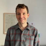 A smiling man in a plaid shirt standing indoors.