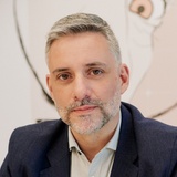 A portrait of a middle-aged man with a trimmed beard, wearing a suit and sitting in front of an abstract artwork.