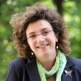 A portrait of a smiling woman with curly brown hair, wearing glasses, a black jacket, and a light green scarf.