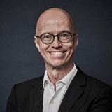 A portrait of a smiling bald man wearina glasses, a dark suit, and a light blue shirt.
