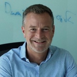 A smiling man in a blue checkered shirt sitting in an office environment.