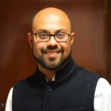 A portrait of a smiling man wearing glasses and a black Nehru jacket over a white shirt.