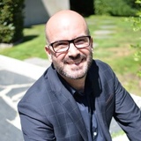 A smiling bald man wearing glasses and a dark blazer is outdoors on a sunny day.