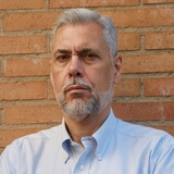 A portrait of a middle-aged man with a serious expression, having gray hair and beard, wearing a light blue shirt against a brick wall background.