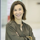 A woman smiling in a professional setting, wearing a khaki blouse and standing with arms crossed.