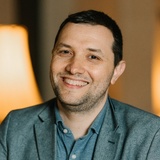 A smiling man wearing a blazer over a shirt and seated indoors.