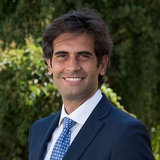 A smiling man in a blue suit and patterned tie stands outdoors with greenery in the background.