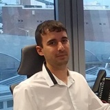 A man in a white shirt sits at an office desk with a city view in the background.