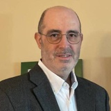 A portrait of a bald man with glasses wearing a grey blazer and a white shirt