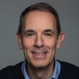 A portrait of a middle-aged man smiling at the camera, wearing a dark sweater and a light blue shirt.