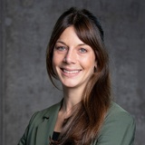 Portrait of a smiling woman with long brown hair wearing a green blazer, posing against a gray background.