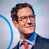 A professional man with glasses is smiling during a public speaking event.