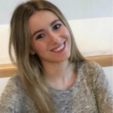 A portrait of a smiling young woman with long blonde hair, wearing a grey sweater, sitting indoors.