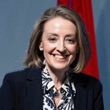 A smiling woman speaking at a podium with a microphone, dressed in a suit with a patterned blouse.