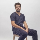A man in a dark blue scrub suit sitting on a wooden stool against a grey background
