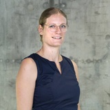 A professional portrait of a woman with glasses, wearing a navy blue sleeveless top, standing in front of a textured concrete background.