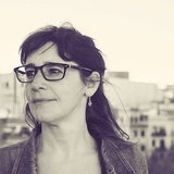 A black and white portrait of a woman with glasses looking away from the camera, standing in front of a cityscape.