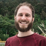 A smiling man with a beard wearing a red shirt, standing outdoors with trees in the background.