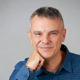 A portrait of a smiling middle-aged man with gray hair, wearing a blue patterned shirt and resting his chin on his hand.