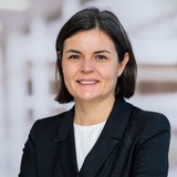 A professional portrait of a smiling woman with short, dark hair, wearing a black blazer over a white top.