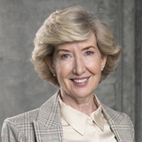 A portrait of an elderly woman with a warm smile, dressed in a professional beige blazer and a light beige blouse.