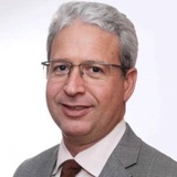 A portrait of a smiling middle-aged man with silver hair, wearing glasses, a gray suit, and a brown tie.