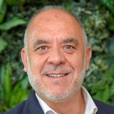 A portrait of a smiling middle-aged man with a beard, wearing a suit jacket over a white shirt, standing outdoors with greenery in the background.