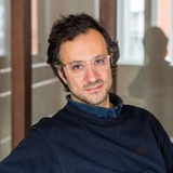 A man with curly hair wearing glasses and a dark blue sweater is seated in a corridor, looking at the camera.