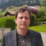 A man in a plaid blazer standing in front of mountains and houses