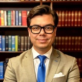 A professional man wearing glasses, a beige suit, and a blue tie is smiling in front of a bookshelf.