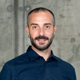 A man with a beard and earrings, wearing a blue shirt, smiling at the camera with a concrete background.