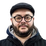 A man with a beard, wearing glasses and a black cap, looks directly at the camera.