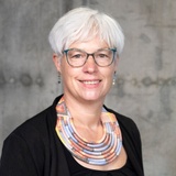 A portrait of a smiling woman with short white hair, wearing glasses and a colorful necklace.