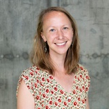 A smiling woman with short blonde hair, wearing a floral dress, standing against a gray concrete background.