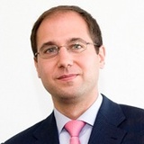 A professional portrait of a man wearing glasses, a dark suit, blue shirt, and pink tie.