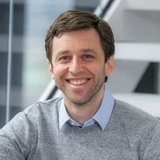 A smiling man with dark hair wearing a grey sweater over a blue shirt in an office setting.