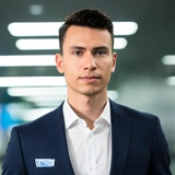 A professional young man in a suit stands confidently in a modern office environment.