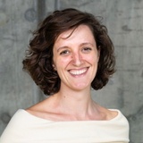 A portrait of a smiling woman with short brown hair, wearing an off-shoulder cream top against a textured gray background.