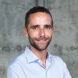 A professional portrait of a smiling man with a beard, wearing a blue checked shirt.
