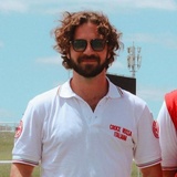 A man with curly hair wearing sunglasses and a white polo shirt with 'Croce Rossa Italiana' embroidered on it stands outdoors.