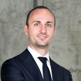 A professional portrait of a bald man wearing a dark suit with a tie.