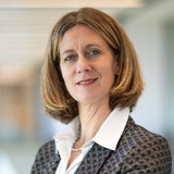 A professional woman with short hair, wearing a blazer and blouse, stands in an office setting.