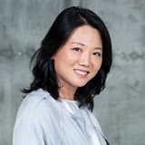 A smiling Asian woman in a light gray blouse against a textured gray backdrop.