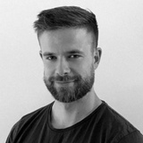 A portrait of a man with a beard, smiling subtly at the camera, wearing a black t-shirt.