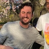 A man with a beard smiling outdoors with cherry blossoms in the background