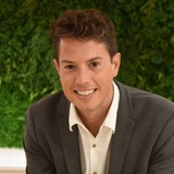 A smiling man in a gray blazer and white shirt sitting in front of a green leafy background.